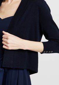 Žena v tmavě modrém cardiganovém svetru s očky na rukávech, pod kterým má odpovídající šaty s plisovanou sukní.