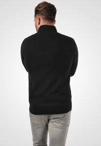 Schwarzer gestrickter Pullover mit einem hohen Kragen, strukturierter Oberfläche und geripptem Saum, kombiniert mit hellgrauen Jeans, die subtile Details aufweisen.