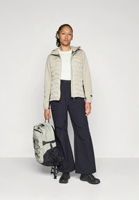 Helebeežist beež värvi punnis jope kapuutsiga, valge särk ja mustad laiad püksid. Tume seljakott hallide detailide ja rihmadega. Mustad kingad.