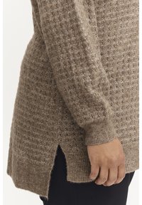 Un long pull en tricot lâche de couleur beige chaud avec une texture en tissage ouvert, des poignets côtelés et des fentes sur les côtés en bas.