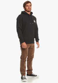 Felpa nera con tasca a marsupio e logo sul lato sinistro del petto, abbinata a pantaloni in cotone a coste marroni e sneakers nere. Vestibilità casual, misto cotone.