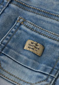 Lys blå denimstoff med en liten teksturert etikett sydd på en lomme, med teksten "BETTER TOGETHER NAME IT."