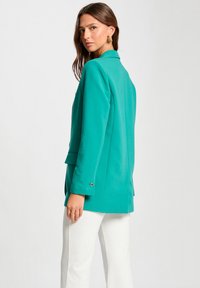 Blazer azul verdoso hecho de una tela texturizada, con un cuello con muescas, puños de un solo botón y bolsillos laterales, estilizado sobre pantalones blancos.