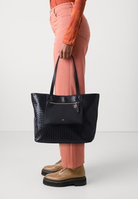 Czarna torba typu tote o plecionej fakturze, z przednią kieszenią na zamek i złotymi ozdobami. Dobrania do jasnoróżowych spodni i brązowych sznurowanych butów.