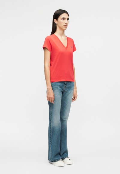 Γυναίκα που στέκεται πλάγια, φοράει κόκκινη μπλούζα με V λαιμόκοψη, μπλε τζιν καμπάνα και λευκά αθλητικά παπούτσια, σε απλό λευκό φόντο.