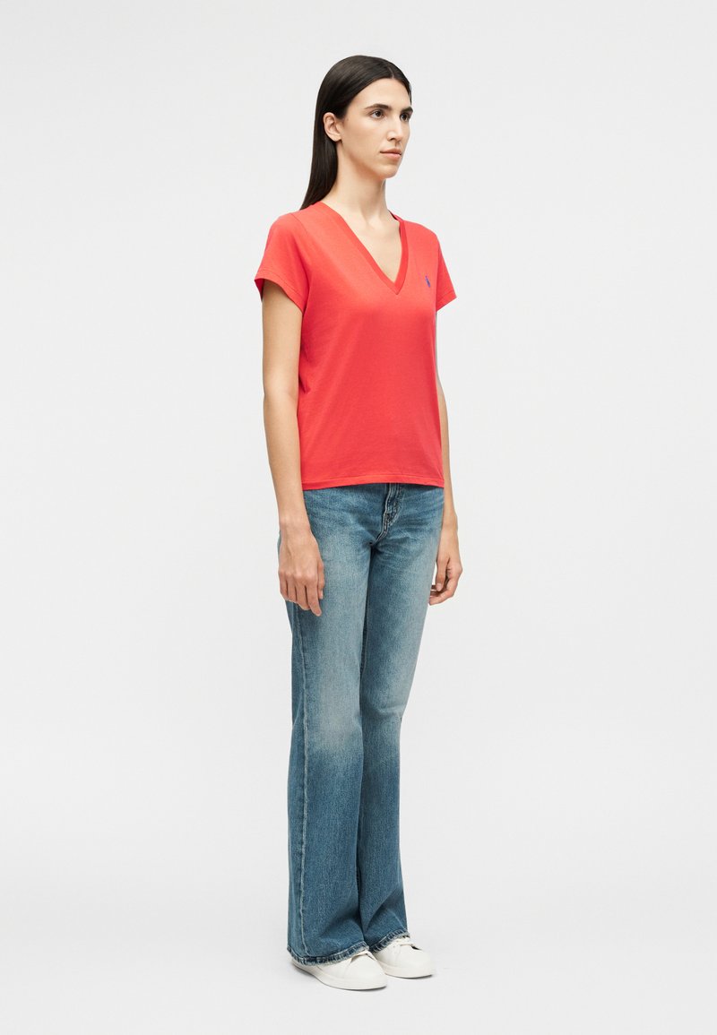 Vrouw staand opzij, gekleed in een rood T-shirt met V-hals, blauwe bootcut jeans en witte sneakers tegen een effen witte achtergrond.