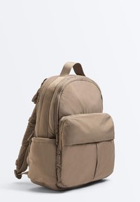Beiger gepolsterter Rucksack mit vorderer Reißverschlusstasche, Seitentaschen und verstellbaren Schulterriemen auf neutralem hellem Hintergrund.