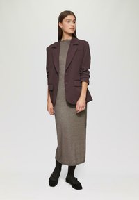 Bordowy żakiet z poduszkami na ramionach i dwoma guzikami, zestawiony z dopasowaną sukienką z metalicznej dzianiny. Modelka nosi czarne rajstopy i miękkie czarne buty.