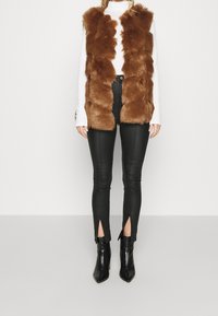Gilet in finta pelliccia marrone con una lunga texture shaggy sopra una maglietta bianca a maniche lunghe, abbinato a pantaloni neri attillati con fessure alla caviglia e stivali.