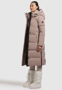 Geacă puffer bej deschis, lungime până la genunchi, cu glugă și buzunare cu fermoar. Jacheta are un model matlasat și accente negre.