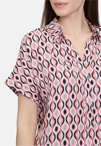 Blusa a maniche corte realizzata in tessuto leggero, caratterizzata da un motivo ondulato nei colori rosa, nero e bianco con forme ovali e una patta con bottoni.