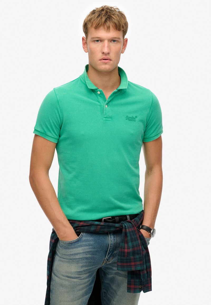 Camisa polo verde de algodón, con cuello clásico, corte ajustado y un pequeño logo bordado. Combinada con jeans azules y una camisa a cuadros atada a la cintura.