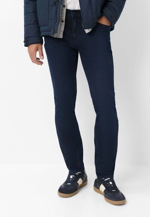 Mann, der dunkelblaue Jeans, marineblaue Sneakers mit weißen Streifen, eine blaue Jacke und ein kariertes Hemd mit einem weißen T-Shirt darunter trägt.