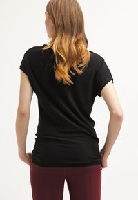 T-shirt noir à manches courtes en tissu léger, présentant une silhouette ajustée et une texture lisse. Vue de dos montrant de longs cheveux en couches.