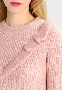 Femme blonde portant un pull rose pâle en tricot avec un détail froncé sur la poitrine et l'épaule, lèvres peintes en rose vif.