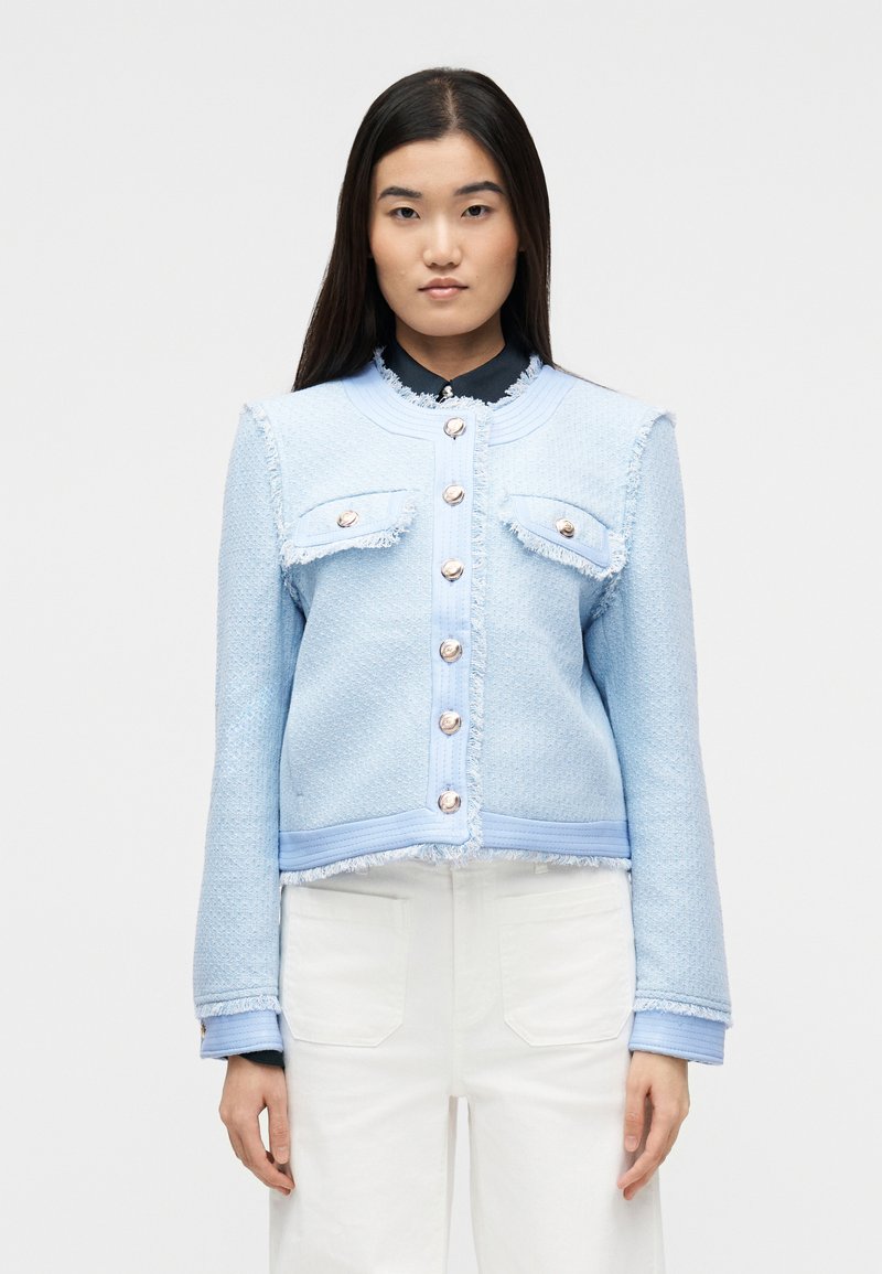 Νεαρή γυναίκα που φοράει ένα ανοιχ τό μπλε υφασμάτινο σακάκι με χρυσά κουμπιά και άσπρο παντελόνι, στέκεται μπροστά σε ένα απλό λευκό φόντο.