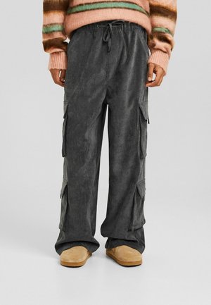 Person iført løse mørkegrå cargobukser med sidelommer, en ferskenstribet sweater og brune ruskindssko, stående på en ensfarvet baggrund.