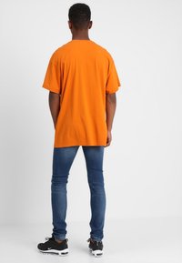Camiseta de algodón naranja con un ajuste casual, combinada con jeans ajustados de color azul y zapatillas negras con acentos blancos. El fondo es liso.