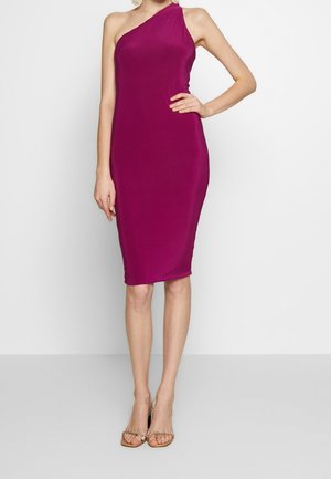 Vrouw die een knielange, asymmetrische magenta jurk draagt en open schoenen met juwelenbandjes, staand tegen een effen achtergrond.