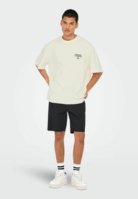 Camiseta oversized color crema con estampado gráfico, combinada con pantalones cortos negros texturizados y zapatillas blancas con acentos negros.