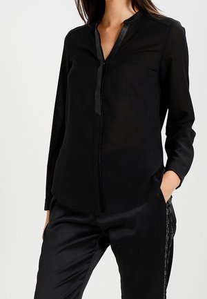 Schwarze Damenbluse mit langen Ärmeln und verdeckten Knöpfen, dazu passende schwarze Hosen mit Seitenstreifen, Hände in den Taschen.