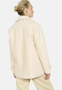 Beżowa, oversize'owa kurtka o miękkiej fakturze, z dużym kołnierzem i mankietami na guziki, zestawiona z kremowymi, prążkowanymi spodniami.