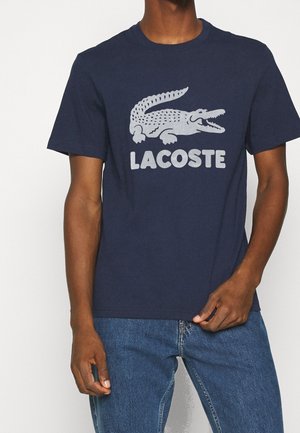Mężczyzna ubrany w granatowy t-shirt Lacoste z dużym białym logo krokodyla i napisem, w połączeniu z niebieskimi dżinsami.