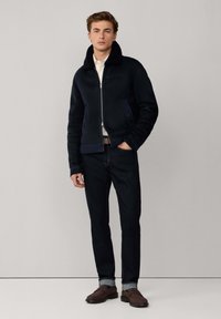 Joven vestido con una chaqueta de piel negra, jeans oscuros con puños, mocasines marrones y una camisa de color claro, de pie contra un fondo gris.