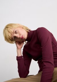 Persona bionda con la testa appoggiata sulla mano, che indossa un dolcevita bordeaux e pantaloni beige, su uno sfondo bianco uniforme.