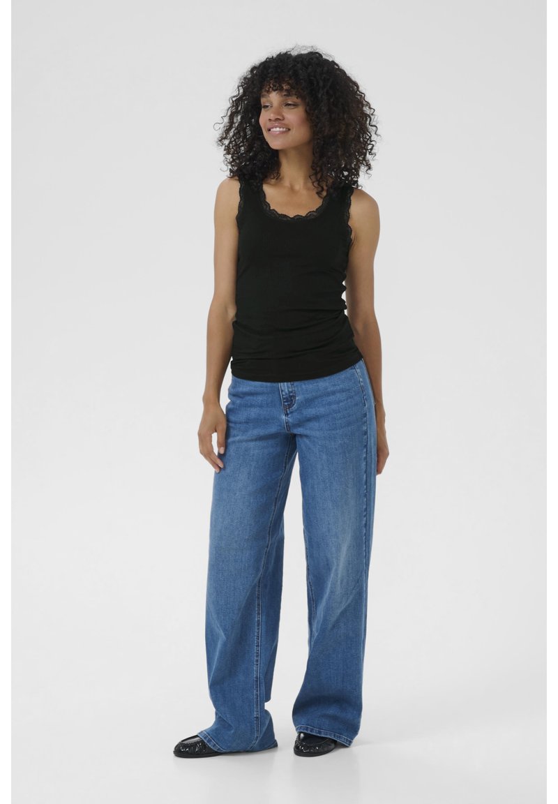 Eine lächelnde Frau mit lockigem Haar steht selbstbewusst in einem schwarzen Tanktop und weiten blauen Jeans vor einem hellgrauen Hintergrund.