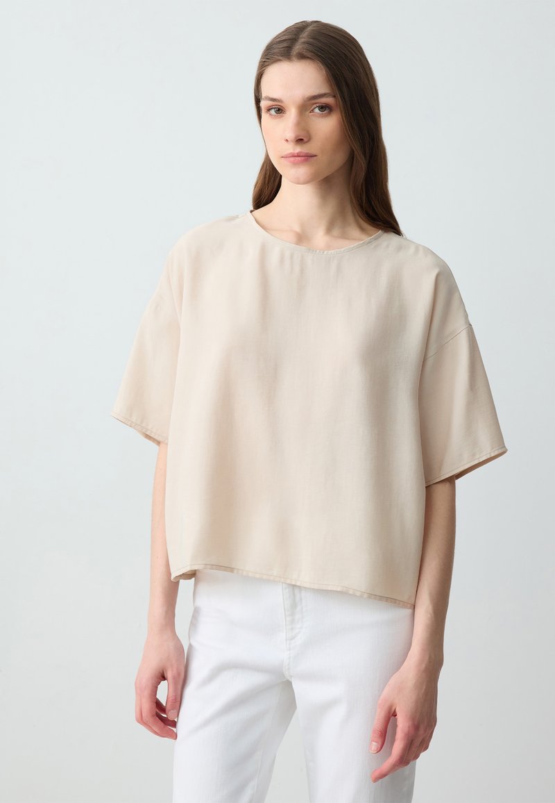Beige blouse met korte mouwen van lichtgewicht stof, met een losse pasvorm en ronde hals. Witte broek eronder voor contrast.