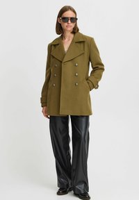 Cappotto doppiopetto verde oliva con un ampio colletto, bottoni argentati e tasche laterali. Abbinato a pantaloni in pelle nera a gamba larga e occhiali da sole scuri.