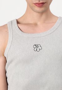 Harmaa ribbattu tankkitoppi, jossa on leveä kaula-aukko ja musta kukkaembroidery-yksityiskohta rinnassa. Pehmeä kangas, joka istuu hyvin vartalon mukaiseen siluettiin.