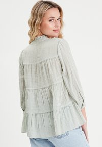 Blouse rayée vert clair avec un ourlet à volants, des manches longues et un col haut. Le tissu semble doux avec une finition texturée.