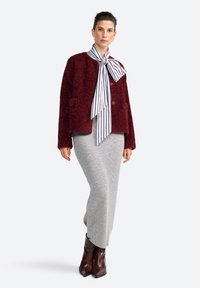 Femme portant une veste texturée bordeaux, un nœud papillon rayé et une jupe côtelée grise, se tenant avec les mains dans les poches et portant des bottes brunes.