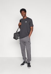 Camiseta deportiva negra con un pequeño logo blanco, combinada con pantalones cargo grises que tienen bolsillos laterales y rayas, y zapatillas negras.