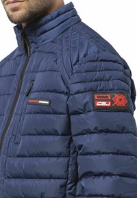 Veste matelassée bleu marine avec détails cousus, fermeture éclair à l'avant et écussons noirs/rouges sur le haut du bras ; tissu lisse et texturé.