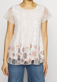 Haut léger et transparent rose clair avec des motifs floraux et circulaires à l'ourlet, doté d'un col rond et d'une fermeture boutonnée sur le devant. Manches courtes volantées.