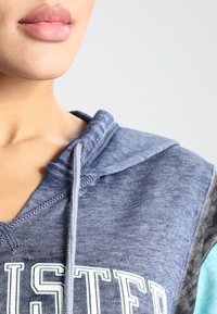 Blå hoodie med en texturerad yta, med en huva som har snörning, stora vita bokstäver på framsidan och färgblockerade ärmar i grått och teal.
