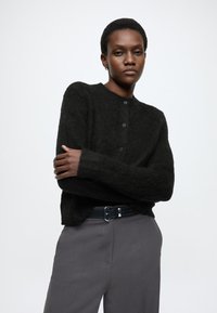 Czarny, teksturowany kardigan z okrągłym dekoltem i pięcioma guzikami, noszony na szarych spodniach z prostym paskiem. Miękki, casualowy design.