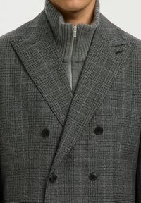 Grå rutad dubbelknäppt kappa med slagkragar, svarta knappar och en grå ribbad zip-up turtleneck under.