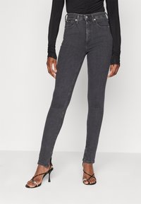 High-waisted black jeans with a slim fit, made of denim. Features ankle slits and subtle stitching detail. Worn with black strappy heels.