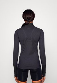 Maglietta sportiva a maniche lunghe di colore grigio scuro con colletto alto. Presenta dettagli riflettenti sul retro e polsini delle maniche neri e testurizzati.