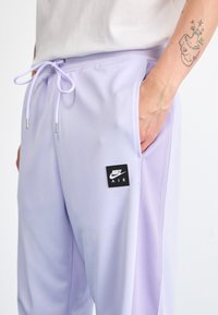 Gaiši lavandas krāsas Nike bikses ar elastīgu jostu, auklu, sānu kabatām un melnu kvadrātveida logotipa uzlīmi, kurā rakstīts "AIR". Gluda tekstūra.