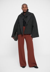 Chaqueta de mezcla de lana negra con un amplio cuello y mangas acampanadas, combinada con pantalones de pierna ancha de color óxido de cintura alta.
