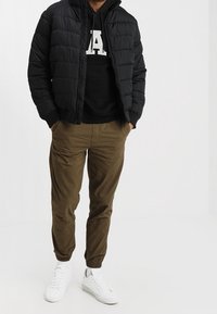 Svart pufferjacka med horisontellt kviltat mönster, kombinerad med en svart hoodie med vit text, och olivgrön tapered pants. Vita sneakers.