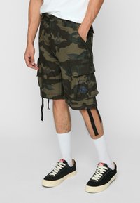 Șorturi cargo cu camuflaj în nuanțe de verde și maro, cu multiple buzunare, curele ajustabile și un patch cu logo albastru. Asortate cu adidași negri și șosete albe.