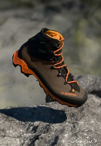 Botă de drumeție maro și portocalie cu suport înalt pentru gleznă, suprafață texturată și talpă din cauciuc. Are șireturi negre și un branding distinct.