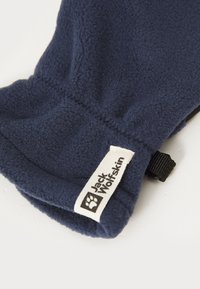 Tumši zili flīsa cimdi ar Jack Wolfskin logo plāksnīti un melnu regulējamu siksniņu uz balta fona.