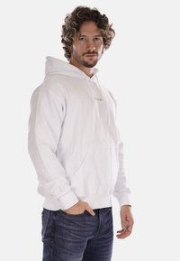 Felpa bianca in cotone con tasca anteriore e cappuccio regolabile; persona in jeans blu appoggiata a uno sfondo semplice.